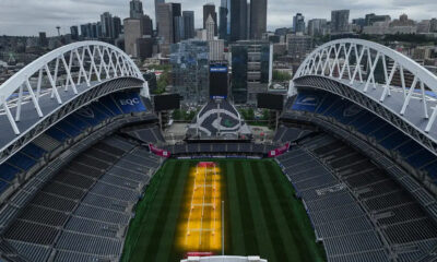 Aerial view of a large empty stadium with white arches, city skyline in the background, and a green field with yellow training lanes in the center.