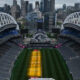 Aerial view of a large empty stadium with white arches, city skyline in the background, and a green field with yellow training lanes in the center.