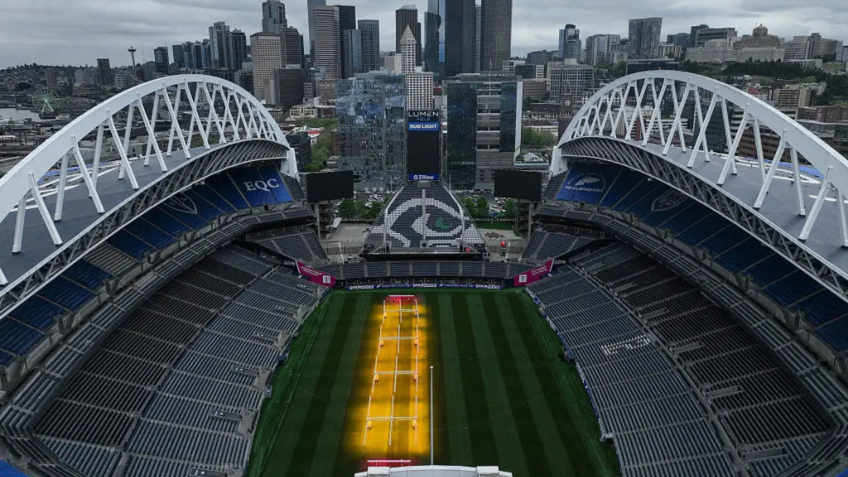 Aerial view of a large empty stadium with white arches, city skyline in the background, and a green field with yellow training lanes in the center.