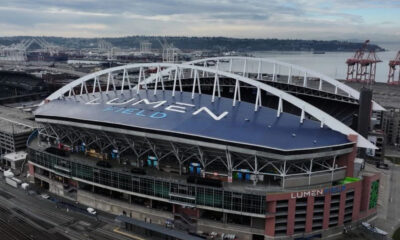 Aerial view of Lumen Field stadium in Seattle, with a blue roof, white arches, and port cranes in the background by the water.
