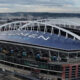 Aerial view of Lumen Field stadium in Seattle, with a blue roof, white arches, and port cranes in the background by the water.