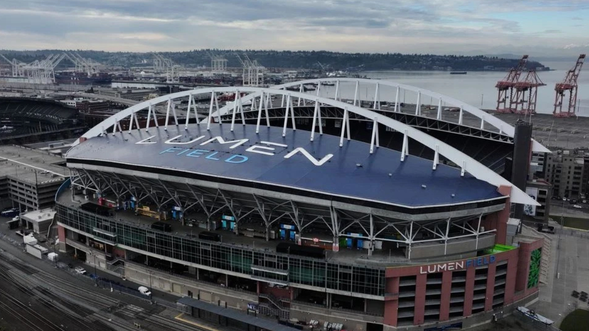 Aerial view of Lumen Field stadium in Seattle, with a blue roof, white arches, and port cranes in the background by the water.