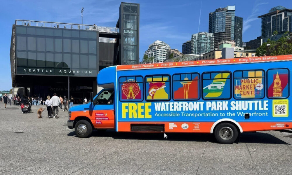 Bright blue and orange shuttle bus with 'FREE WATERFRONT PARK SHUTTLE' lettering parked near Seattle Aquarium, advertising free waterfront transport with windows showing city icons.