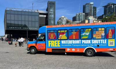 Bright blue and orange shuttle bus with 'FREE WATERFRONT PARK SHUTTLE' lettering parked near Seattle Aquarium, advertising free waterfront transport with windows showing city icons.