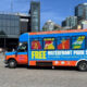 Bright blue and orange shuttle bus with 'FREE WATERFRONT PARK SHUTTLE' lettering parked near Seattle Aquarium, advertising free waterfront transport with windows showing city icons.