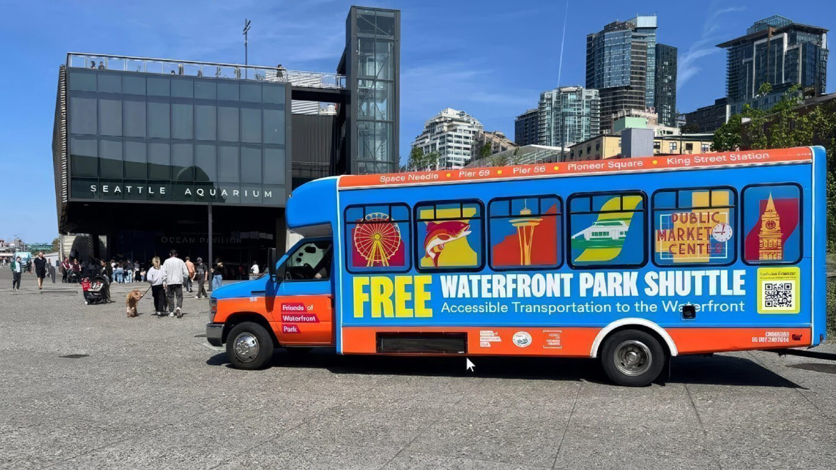 Bright blue and orange shuttle bus with 'FREE WATERFRONT PARK SHUTTLE' lettering parked near Seattle Aquarium, advertising free waterfront transport with windows showing city icons.
