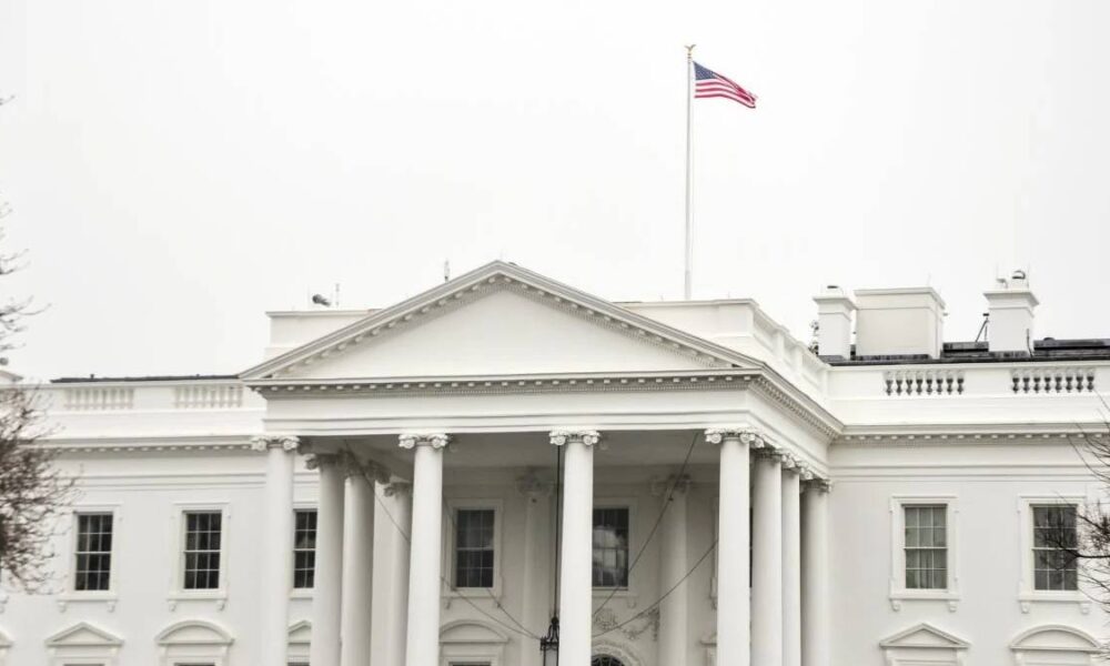 White House facade with tall columns and a flagpole flag flying above the roofline, bright white exterior.