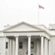 White House facade with tall columns and a flagpole flag flying above the roofline, bright white exterior.