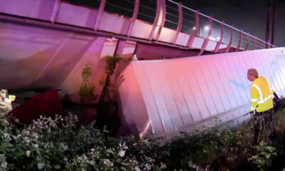 Nighttime scene of a damaged bridge railing and a fallen barrier with a worker in a yellow reflective vest inspecting the site