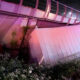 Nighttime scene of a damaged bridge railing and a fallen barrier with a worker in a yellow reflective vest inspecting the site