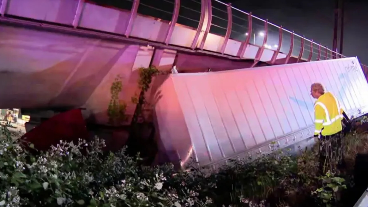 Nighttime scene of a damaged bridge railing and a fallen barrier with a worker in a yellow reflective vest inspecting the site