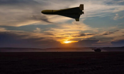 Unmanned military drone flies above a field at sunset; a distant tank sits on the ground.