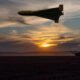 Unmanned military drone flies above a field at sunset; a distant tank sits on the ground.
