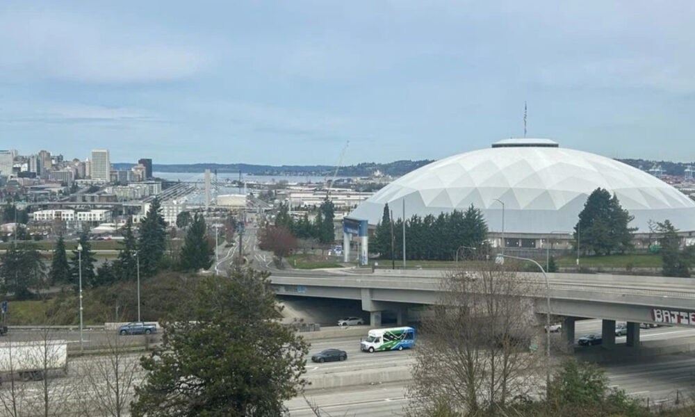 City skyline with a large white geodesic dome stadium beside a highway, trees in the foreground and water in the distance.