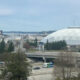 City skyline with a large white geodesic dome stadium beside a highway, trees in the foreground and water in the distance.