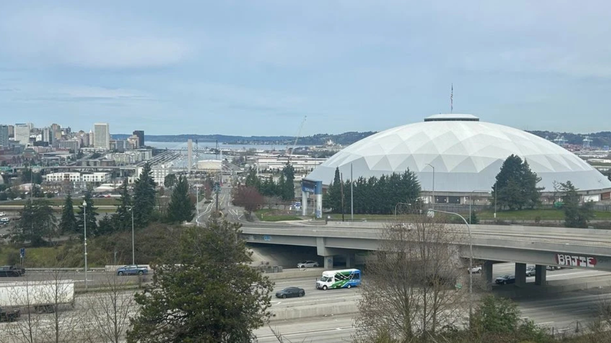 City skyline with a large white geodesic dome stadium beside a highway, trees in the foreground and water in the distance.