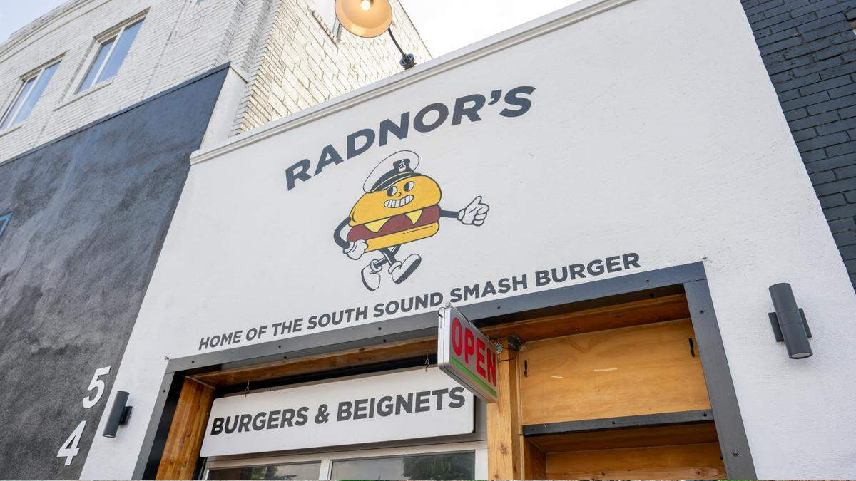 White storefront with sign reading "RADNOR'S" and a cartoon hot dog mascot; below, text says "HOME OF THE SOUTH SOUND SMASH BURGER" and an open sign over the door.
