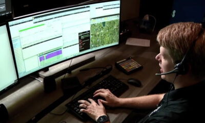 Person wearing a headset types on a keyboard in front of three wide monitors displaying charts and maps, likely monitoring systems.