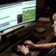 Person wearing a headset types on a keyboard in front of three wide monitors displaying charts and maps, likely monitoring systems.