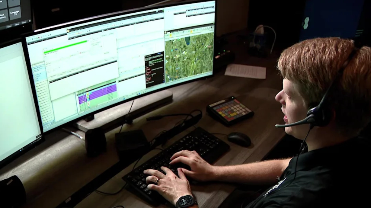 Person wearing a headset types on a keyboard in front of three wide monitors displaying charts and maps, likely monitoring systems.