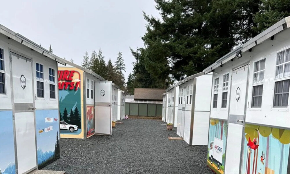 Row of small white cabins with colorful murals on the doors, a gravel path between them and trees in the background.