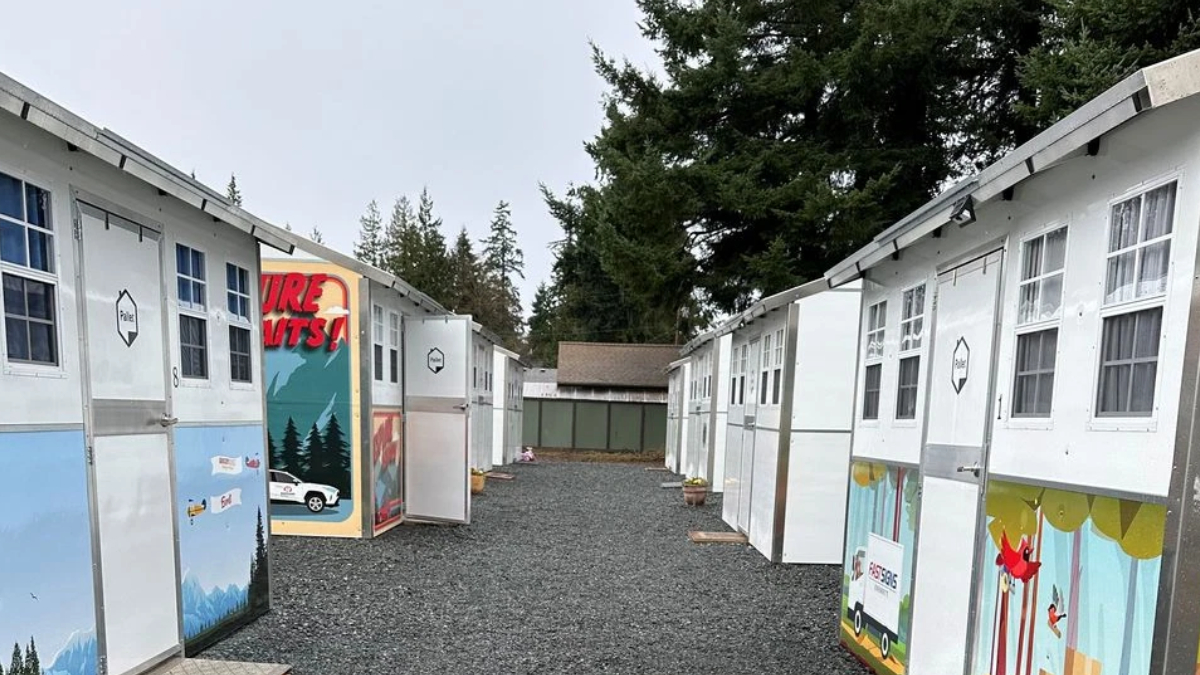 Row of small white cabins with colorful murals on the doors, a gravel path between them and trees in the background.