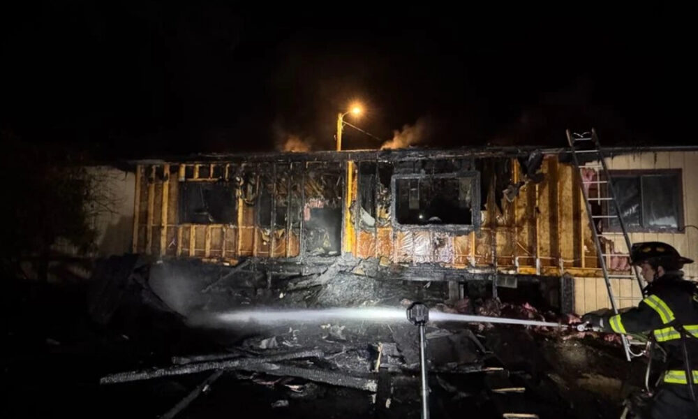 Nighttime scene of a partially burned building with charred walls as a firefighter sprays water on remaining flames from a hose bed.