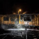Nighttime scene of a partially burned building with charred walls as a firefighter sprays water on remaining flames from a hose bed.