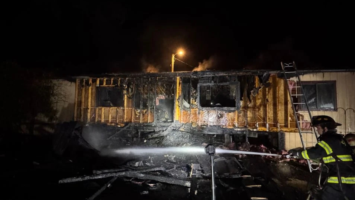 Nighttime scene of a partially burned building with charred walls as a firefighter sprays water on remaining flames from a hose bed.
