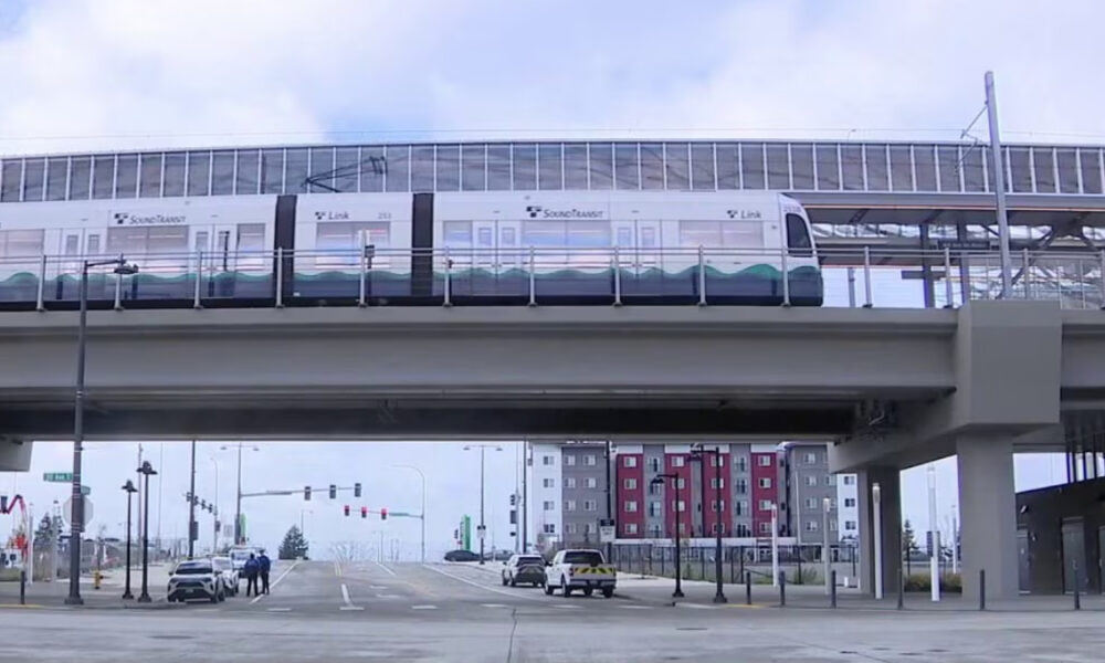 Light rail train runs on an elevated track above a wide urban intersection with cars and pedestrians below. Hazily visible tall apartment buildings in the background.