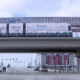 Light rail train runs on an elevated track above a wide urban intersection with cars and pedestrians below. Hazily visible tall apartment buildings in the background.