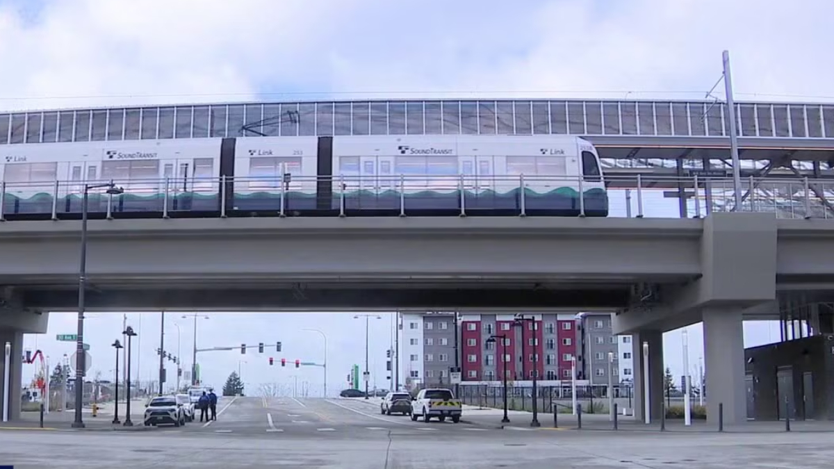 Light rail train runs on an elevated track above a wide urban intersection with cars and pedestrians below. Hazily visible tall apartment buildings in the background.