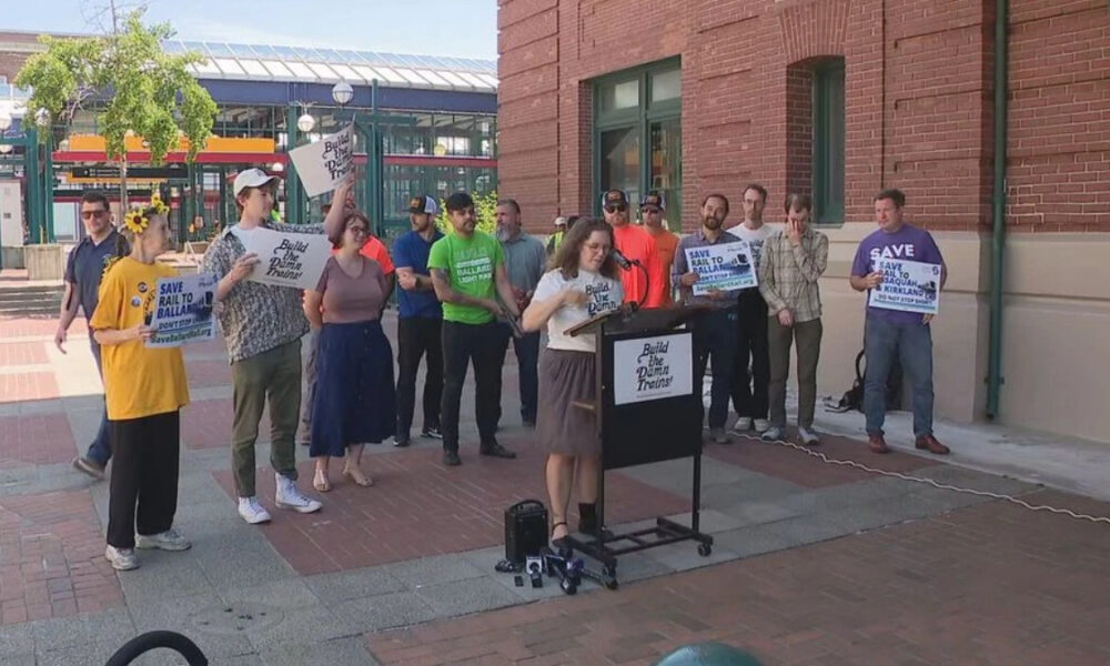 A woman at a mobile podium speaks to a group of demonstrators outside a brick building, holding signs that read 'Build the Damn Trains' and 'Save Rail to Ball...' (activism rally).