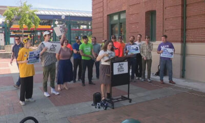 A woman at a mobile podium speaks to a group of demonstrators outside a brick building, holding signs that read 'Build the Damn Trains' and 'Save Rail to Ball...' (activism rally).