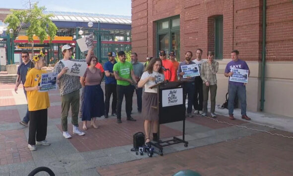 A woman at a mobile podium speaks to a group of demonstrators outside a brick building, holding signs that read 'Build the Damn Trains' and 'Save Rail to Ball...' (activism rally).