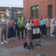 A woman at a mobile podium speaks to a group of demonstrators outside a brick building, holding signs that read 'Build the Damn Trains' and 'Save Rail to Ball...' (activism rally).