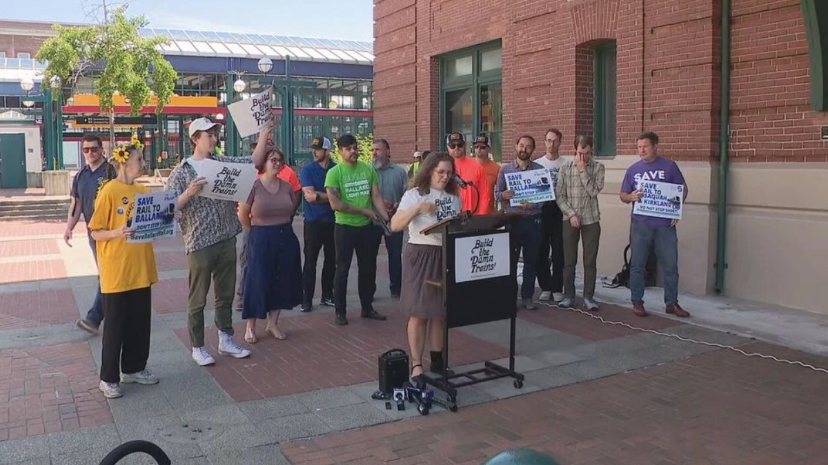 A woman at a mobile podium speaks to a group of demonstrators outside a brick building, holding signs that read 'Build the Damn Trains' and 'Save Rail to Ball...' (activism rally).
