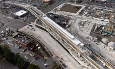 Aerial view of a construction site with an elevated rail line and station under development by roads and parking lots.