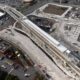 Aerial view of a construction site with an elevated rail line and station under development by roads and parking lots.