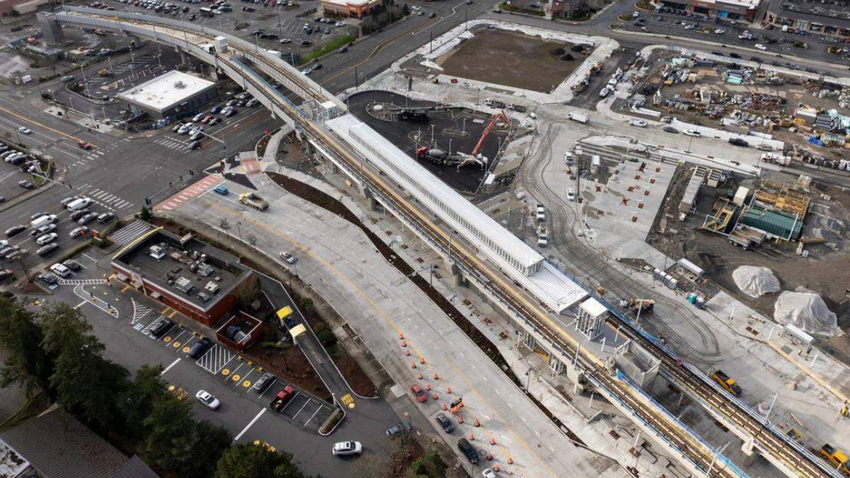 Aerial view of a construction site with an elevated rail line and station under development by roads and parking lots.
