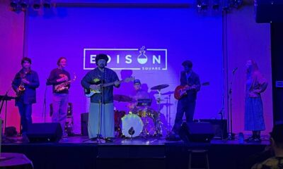 Six-member band performing on stage with blue lighting; guitarist center, drummer in back, others on saxophone and guitar, Edison Square logo on backdrop.