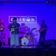 Six-member band performing on stage with blue lighting; guitarist center, drummer in back, others on saxophone and guitar, Edison Square logo on backdrop.