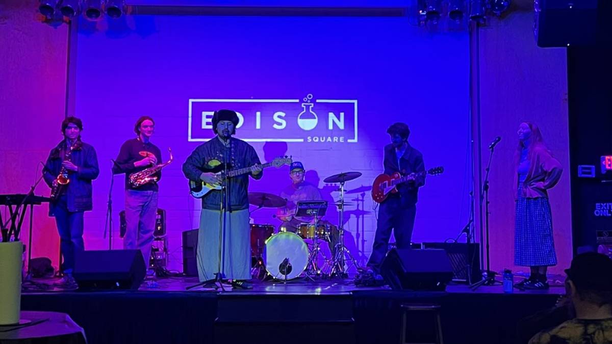 Six-member band performing on stage with blue lighting; guitarist center, drummer in back, others on saxophone and guitar, Edison Square logo on backdrop.