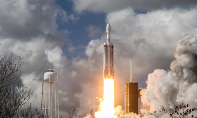 Rocket lifts off from a launch pad with bright orange flame and billowing smoke against a cloudy sky, with support structures and a water tower nearby.