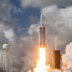Rocket lifts off from a launch pad with bright orange flame and billowing smoke against a cloudy sky, with support structures and a water tower nearby.