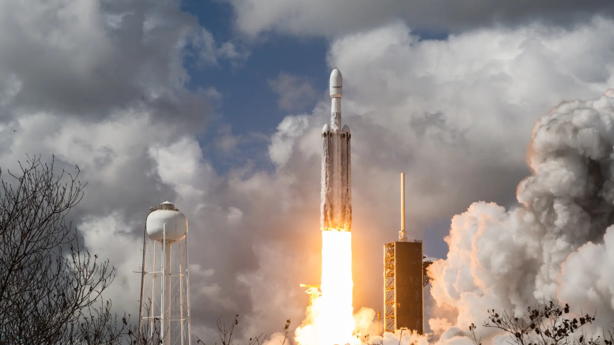 Rocket lifts off from a launch pad with bright orange flame and billowing smoke against a cloudy sky, with support structures and a water tower nearby.