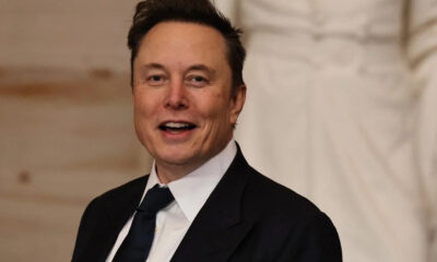Smiling man in a dark suit and tie at a formal event, speaking or presenting.