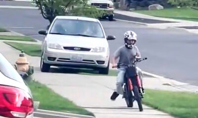 A person wearing a helmet rides a red motorcycle along a residential sidewalk, performing a wheelie with the front wheel lifted off the ground, while a white car is parked on the street nearby.
