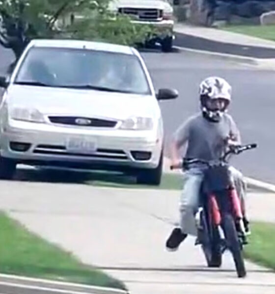 A person wearing a helmet rides a red motorcycle along a residential sidewalk, performing a wheelie with the front wheel lifted off the ground, while a white car is parked on the street nearby.