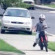 A person wearing a helmet rides a red motorcycle along a residential sidewalk, performing a wheelie with the front wheel lifted off the ground, while a white car is parked on the street nearby.